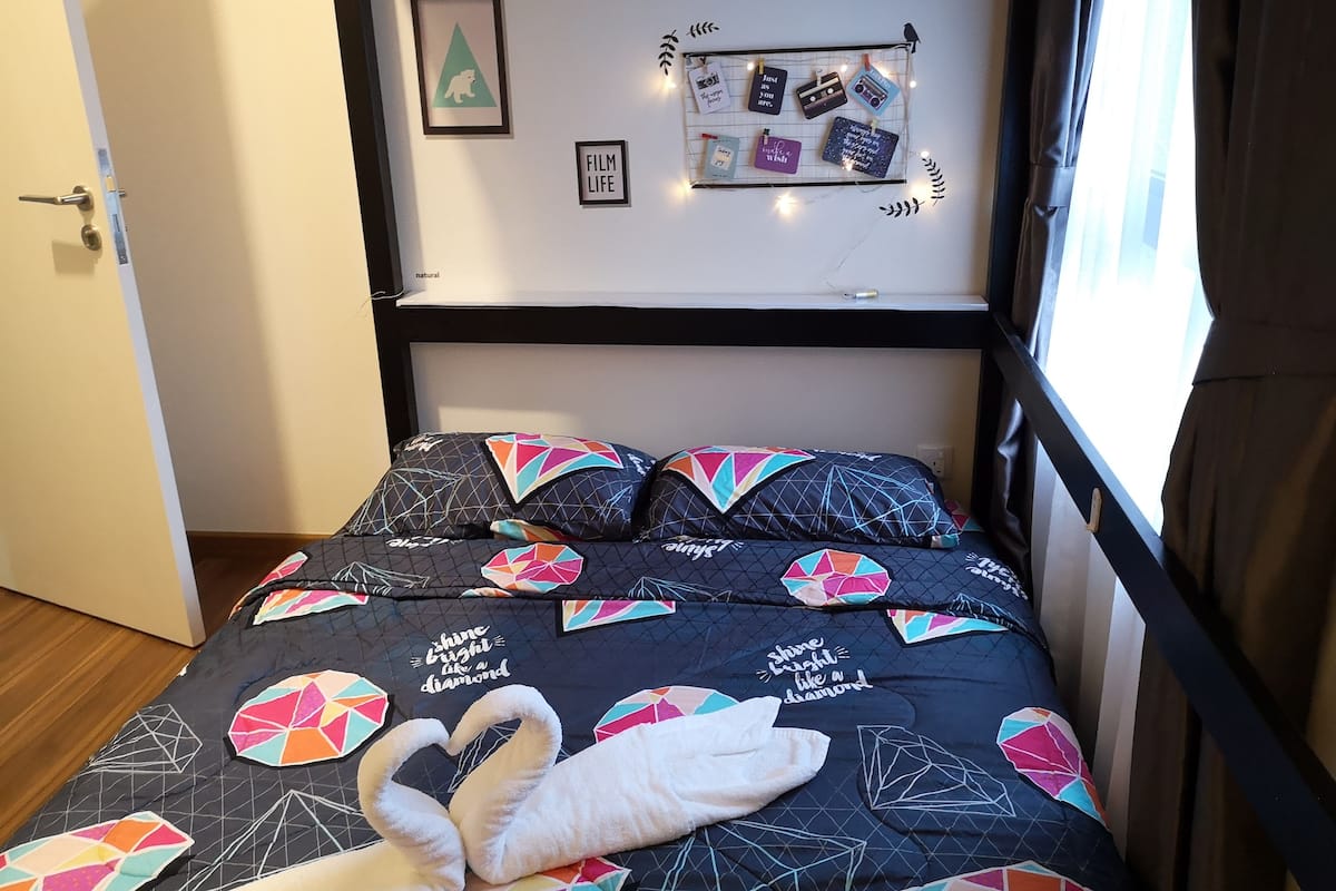 Un lit avec une couette à motifs géométriques, des oreillers et une serviette en forme de cœur. Les murs sont décorés, notamment avec un tableau encadré et une guirlande lumineuse.