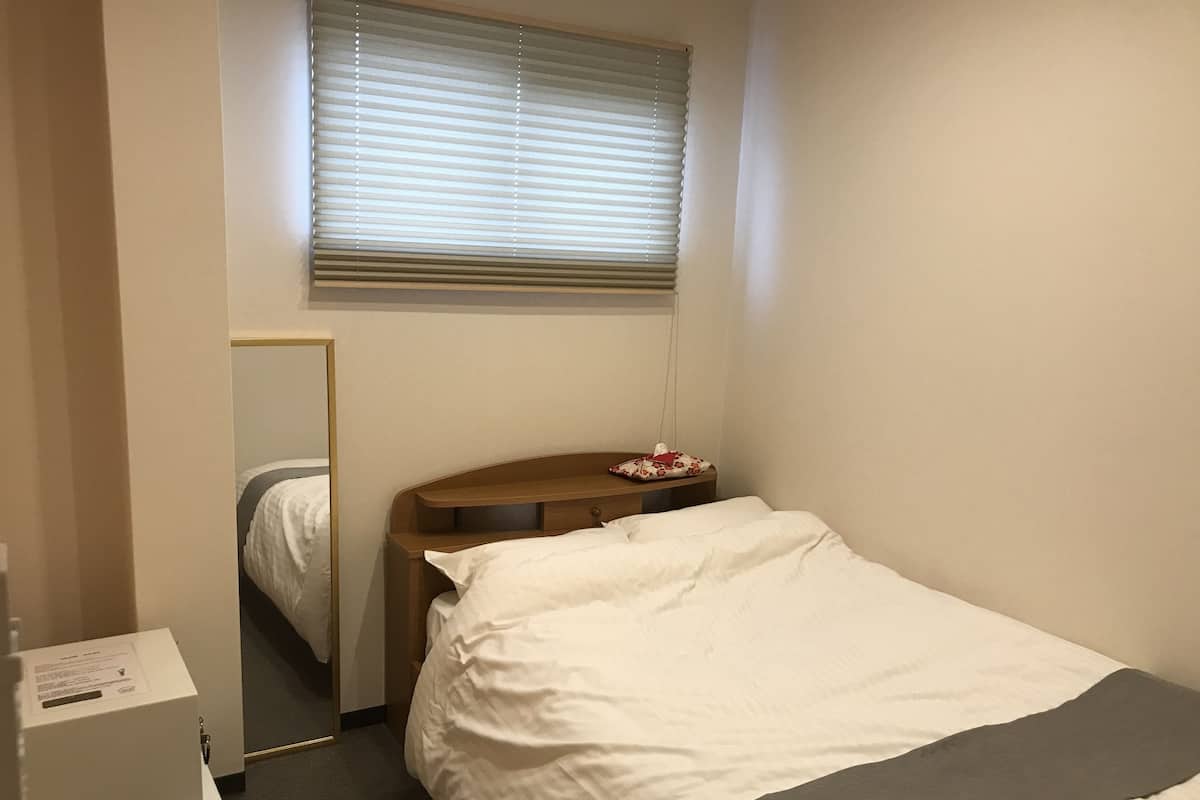 Ein Hotelzimmer mit einem Einzelbett, einem Nachttisch aus Holz, einem Spiegel und einem Fenster mit Jalousien.