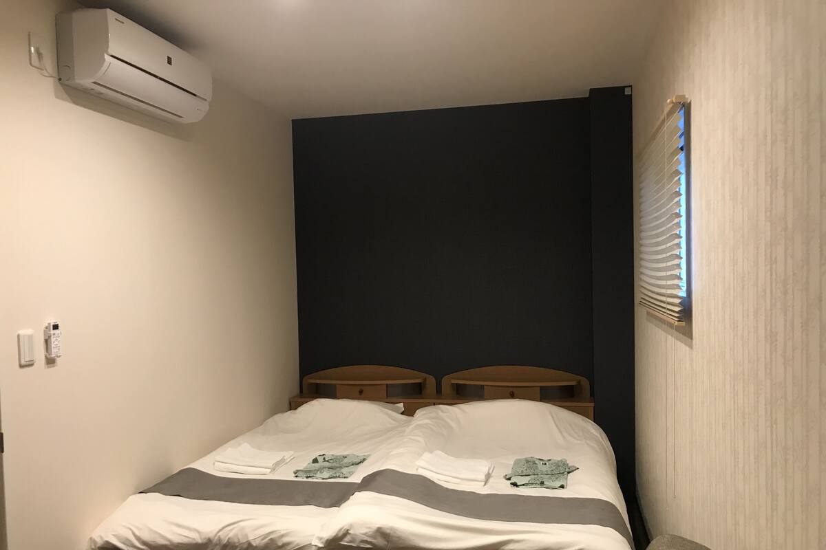 Ein kleines Zimmer mit einem Einzelbett, einer dunklen Akzentwand, einem Fenster mit Jalousien und einer Wand-Klimaanlage.