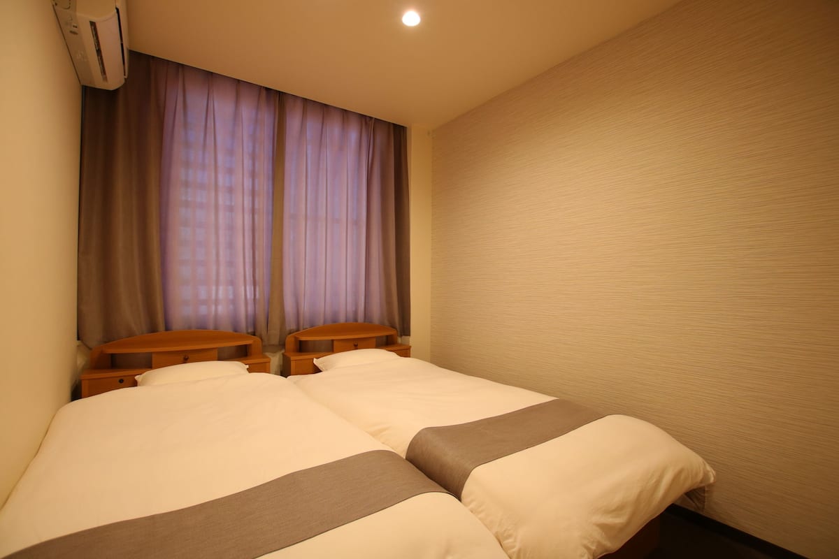 Ein Hotelzimmer mit zwei Betten, einem Holz-Kopfbrett, einer Wand-Klimaanlage und einem Fenster mit Vorhängen.