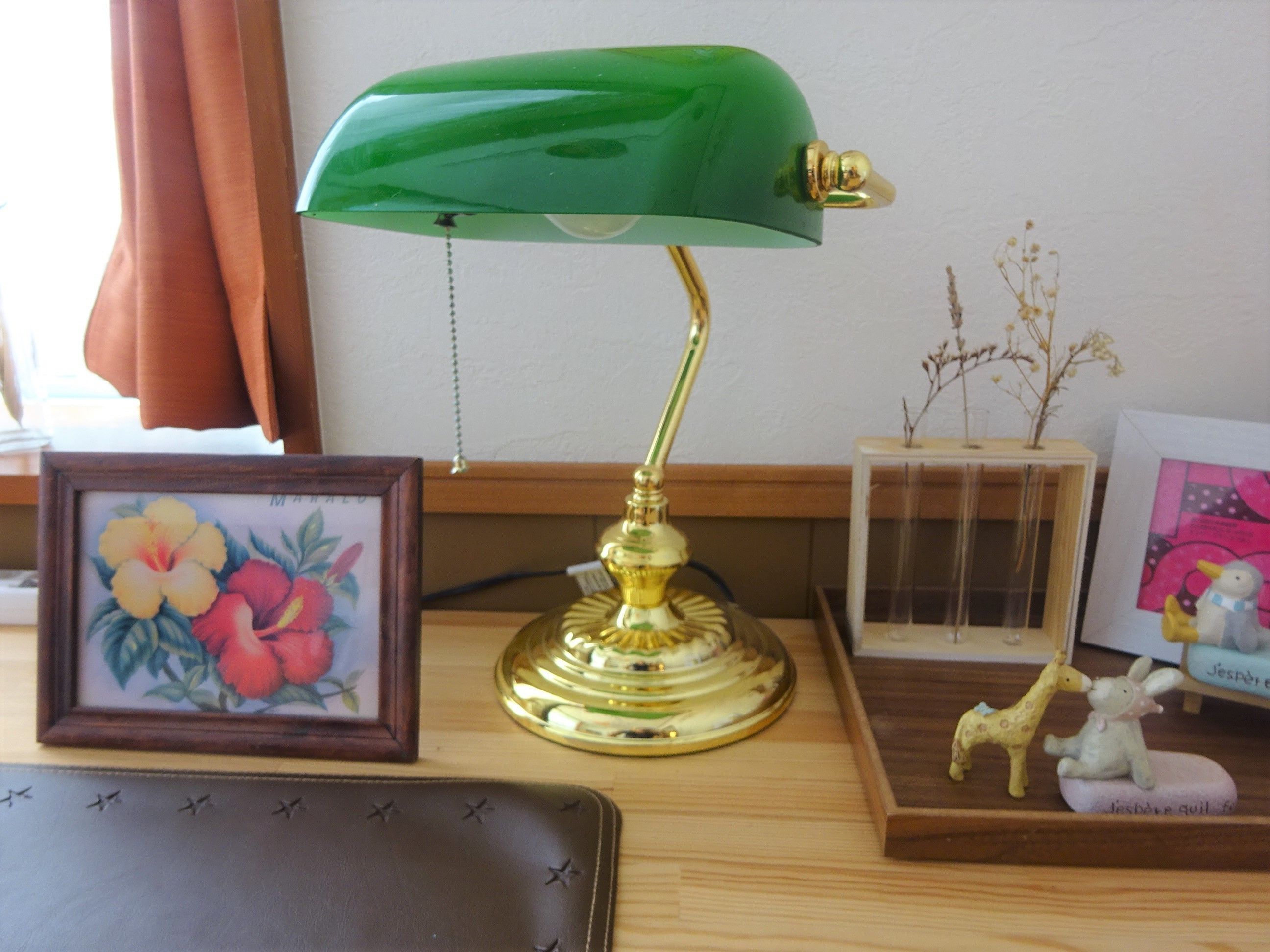 A desk with a green desk lamp, a framed floral picture, a small figurine of a giraffe and a rabbit, and a wooden side table with a picture frame and a small plant.