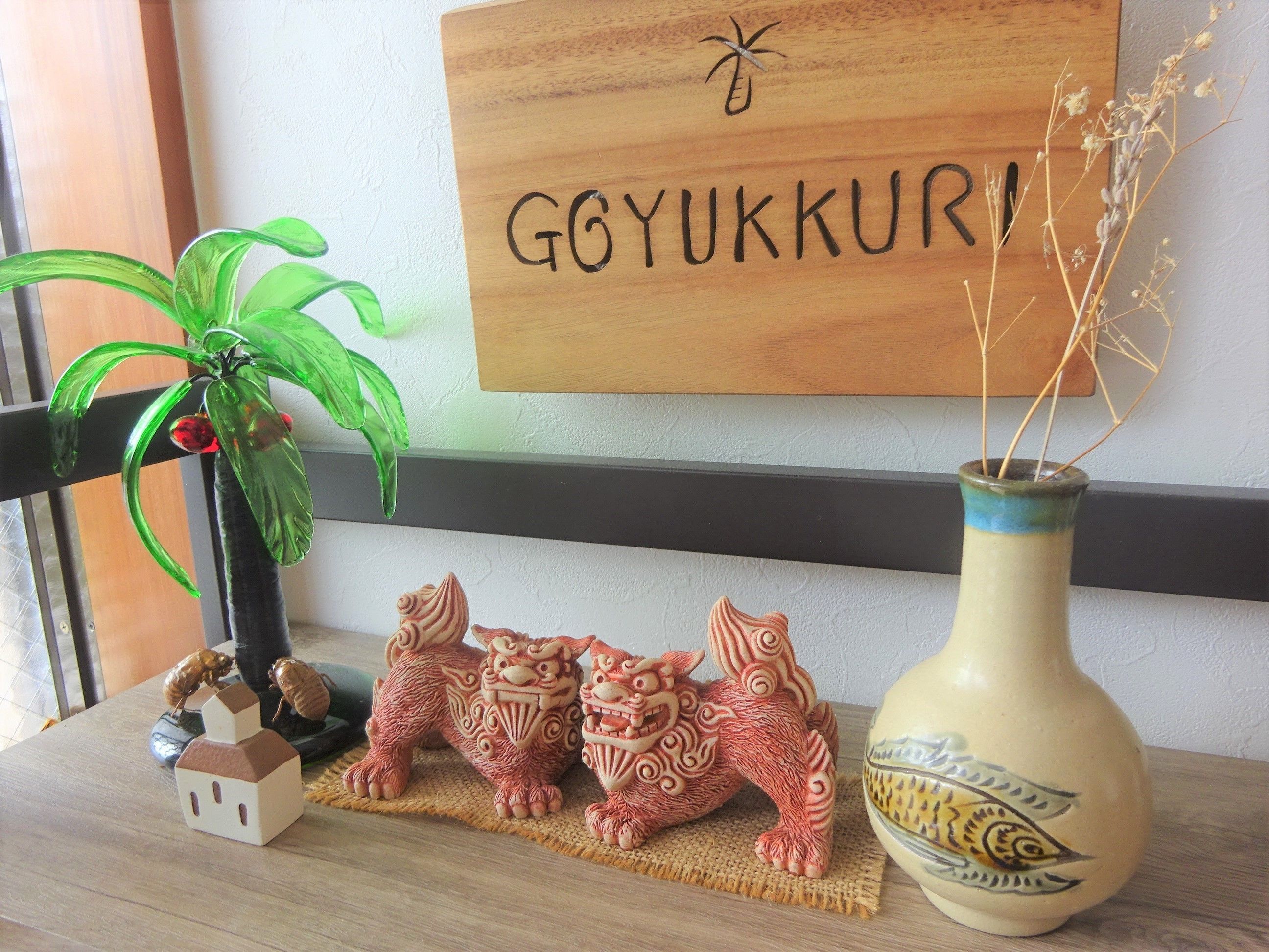 Een houten bord met de tekst 'Goyukkuri' en decoratieve elementen zoals een vaas, een plant en twee leeuwenbeeldjes op een plank.