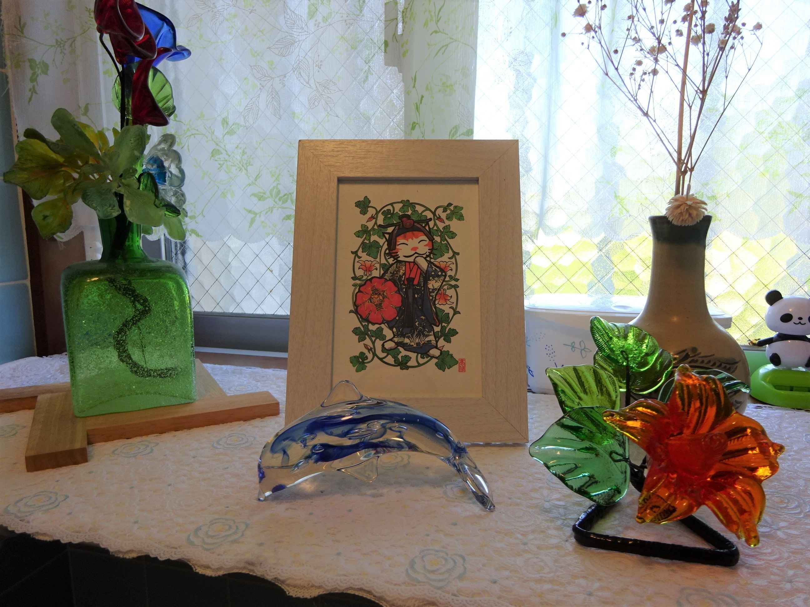 A table with a framed artwork, a green vase with flowers, a blue glass object, and a panda figurine.