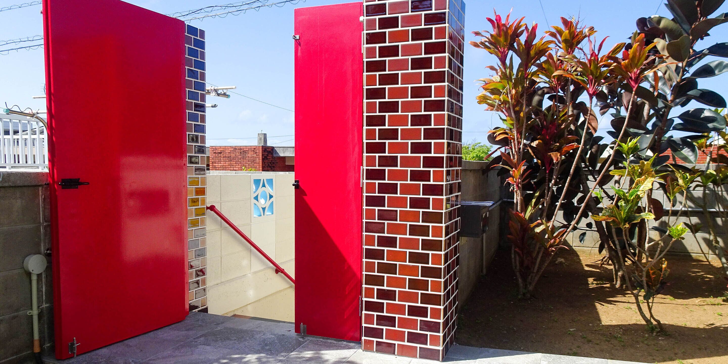 A red gate with a brick pillar, a white fence, and a plant with red leaves.
