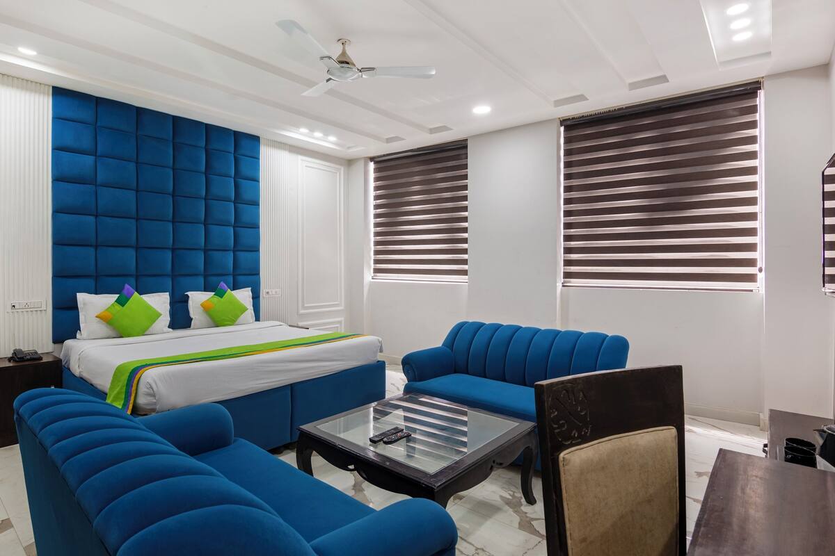 A modern hotel room with a large bed, blue upholstered furniture, a glass coffee table, and a ceiling fan.