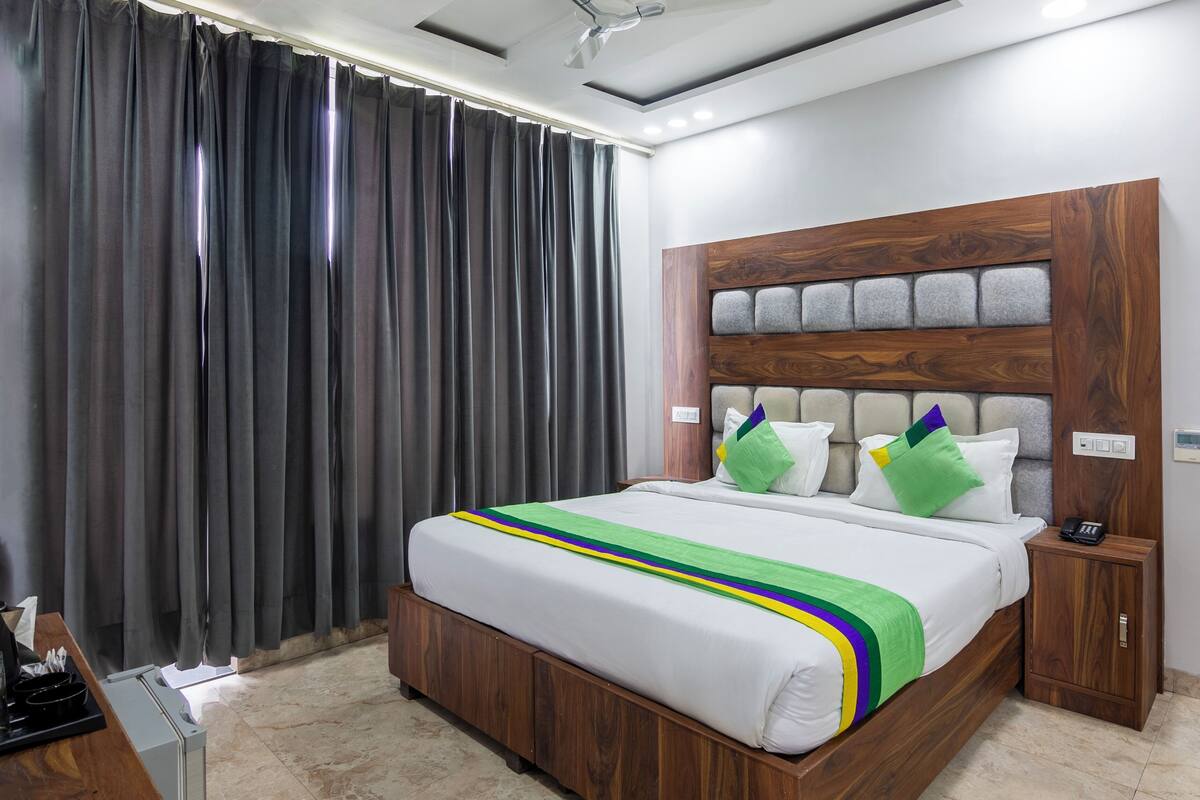 A hotel room with a wooden headboard, a bed with white bedding and a green and yellow striped blanket, a bedside table with a phone, and a wall-mounted thermostat.