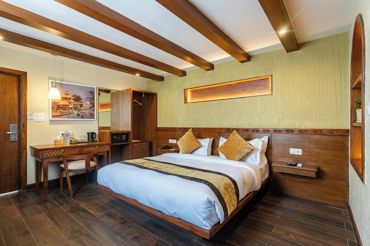 A hotel room with a large bed, wooden headboard, a desk with a chair, a mirror, and a painting of a temple on the wall.