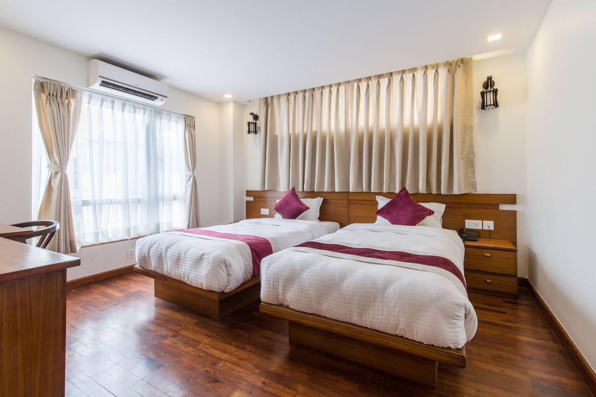 A hotel room with two beds, wooden flooring, a desk, and a window with curtains.