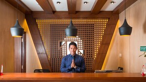 Un mostrador de recepción con detalles de madera, dos lámparas colgantes y un panel decorativo de madera con un diseño geométrico al fondo.