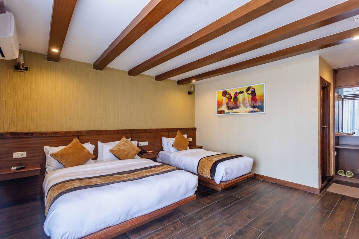 A hotel room with two beds, wooden beams on the ceiling, a painting on the wall, and a shoe rack by the door.