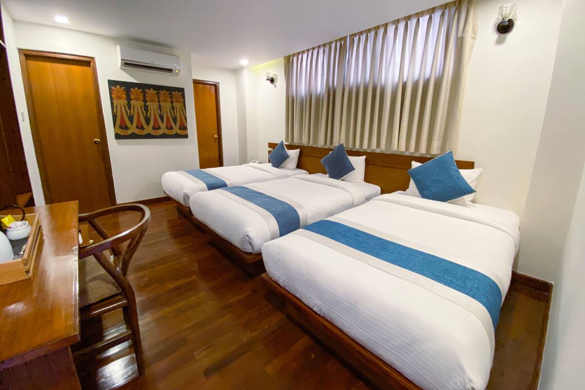 A hotel room with three beds, wooden flooring, a wooden desk, and a painting on the wall.