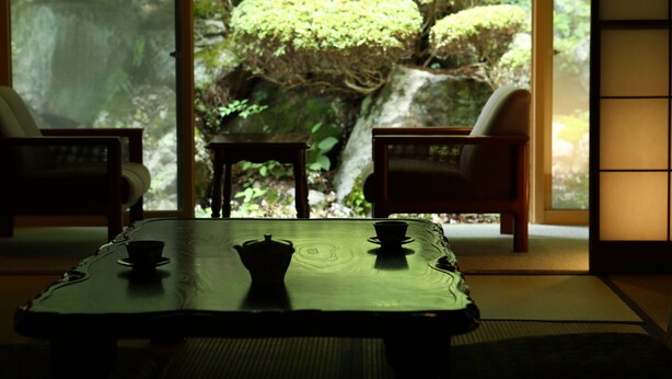 Una habitación japonesa tradicional con suelo de tatami, una mesa baja con un juego de té y vistas a un jardín exuberante a través de amplios ventanales.