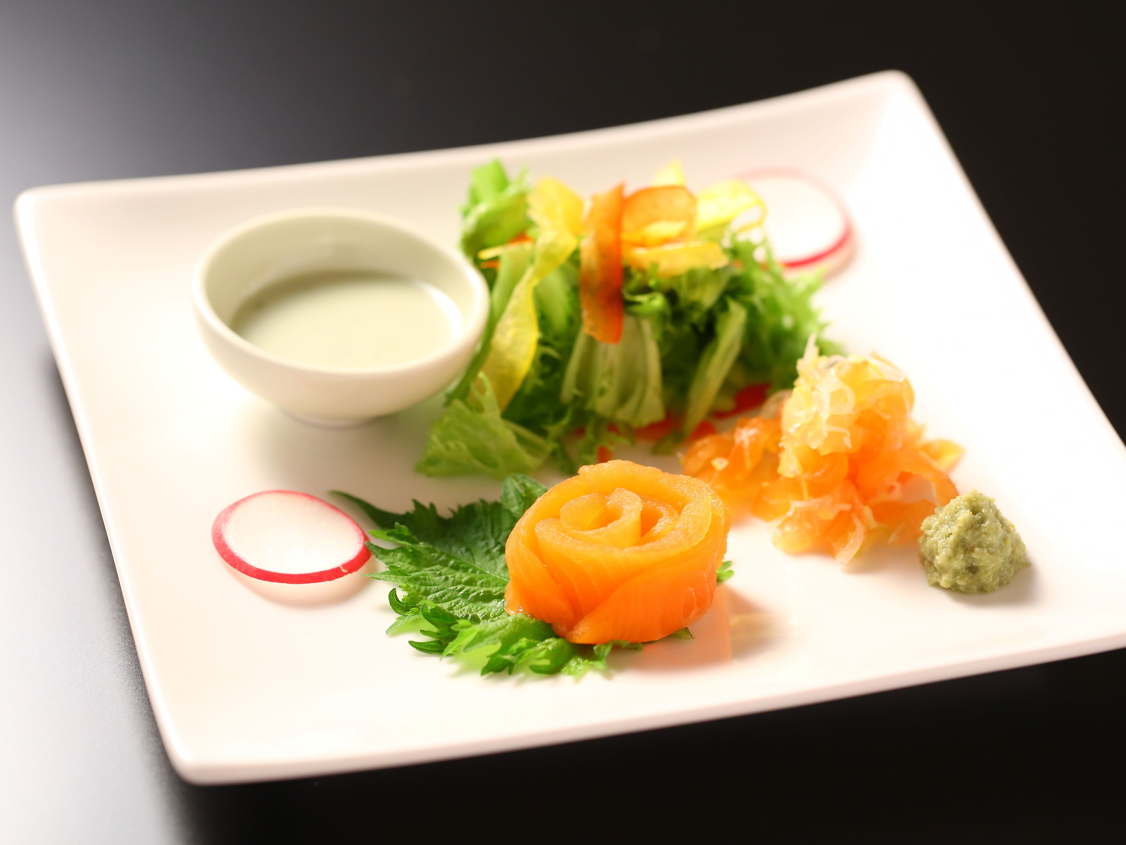 A square plate with a salad, salmon, radish slices, and a small bowl of dressing.