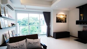 A modern living room with a large window, a sofa with decorative pillows, a black entertainment unit, and a cityscape painting on the wall. - Sabah Luxury Cozy Family Suite (Kota Kinabalu)