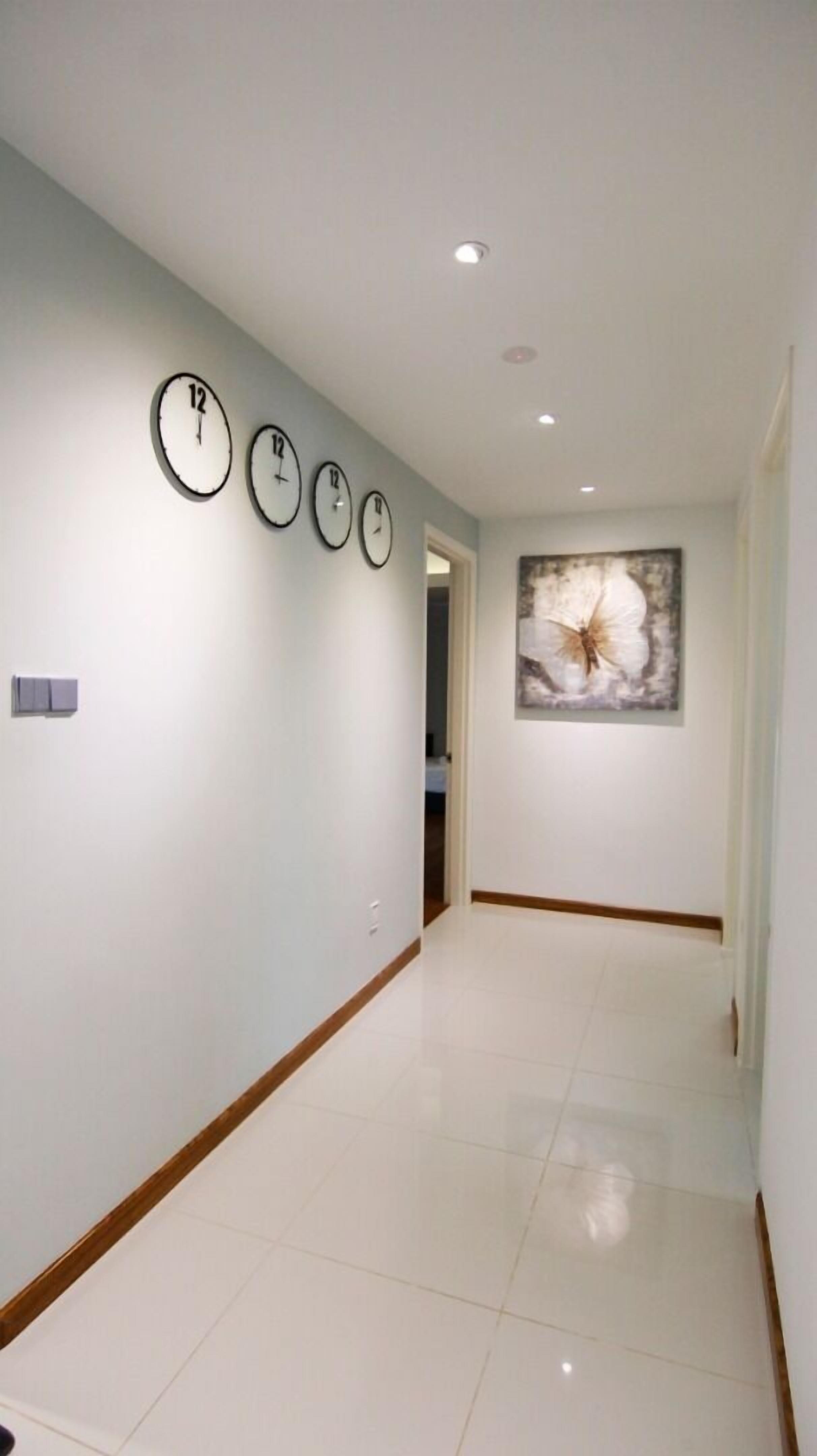 A hallway with white walls, a tiled floor, and four clocks on the left wall.