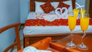 Quarto de hotel com uma cama decorada com pétalas de rosas vermelhas e um enfeite em formato de coração. Duas taças de suco de laranja com canudos e rodelas de abacaxi estão sobre a mesa.