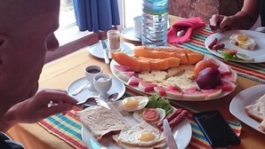 Uma mesa de jantar com diversos alimentos, incluindo frutas fatiadas, ovos e salsichas, em frente a uma parede de pedra e uma janela com cortinas azuis.