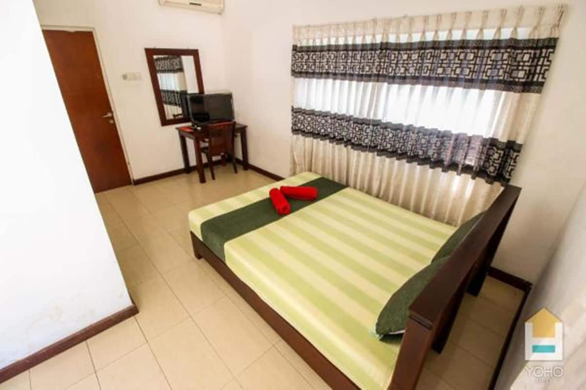 A single bed with a green and white striped bedspread, a red pillow, and a wooden headboard. There is a small wooden table with a television and a mirror on the wall. The room has white walls and a window with patterned curtains.