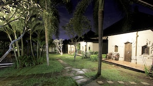 A well-lit courtyard with a stone pathway leading to a building with a traditional design, surrounded by lush greenery and palm trees. - Alamanda (Pecatu)