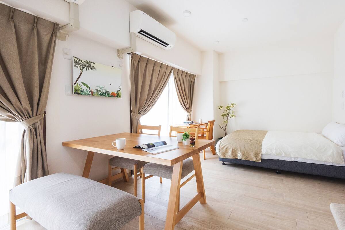 A modern, well-lit room with a wooden dining table, chairs, a bed with a dark headboard, and a small potted plant.