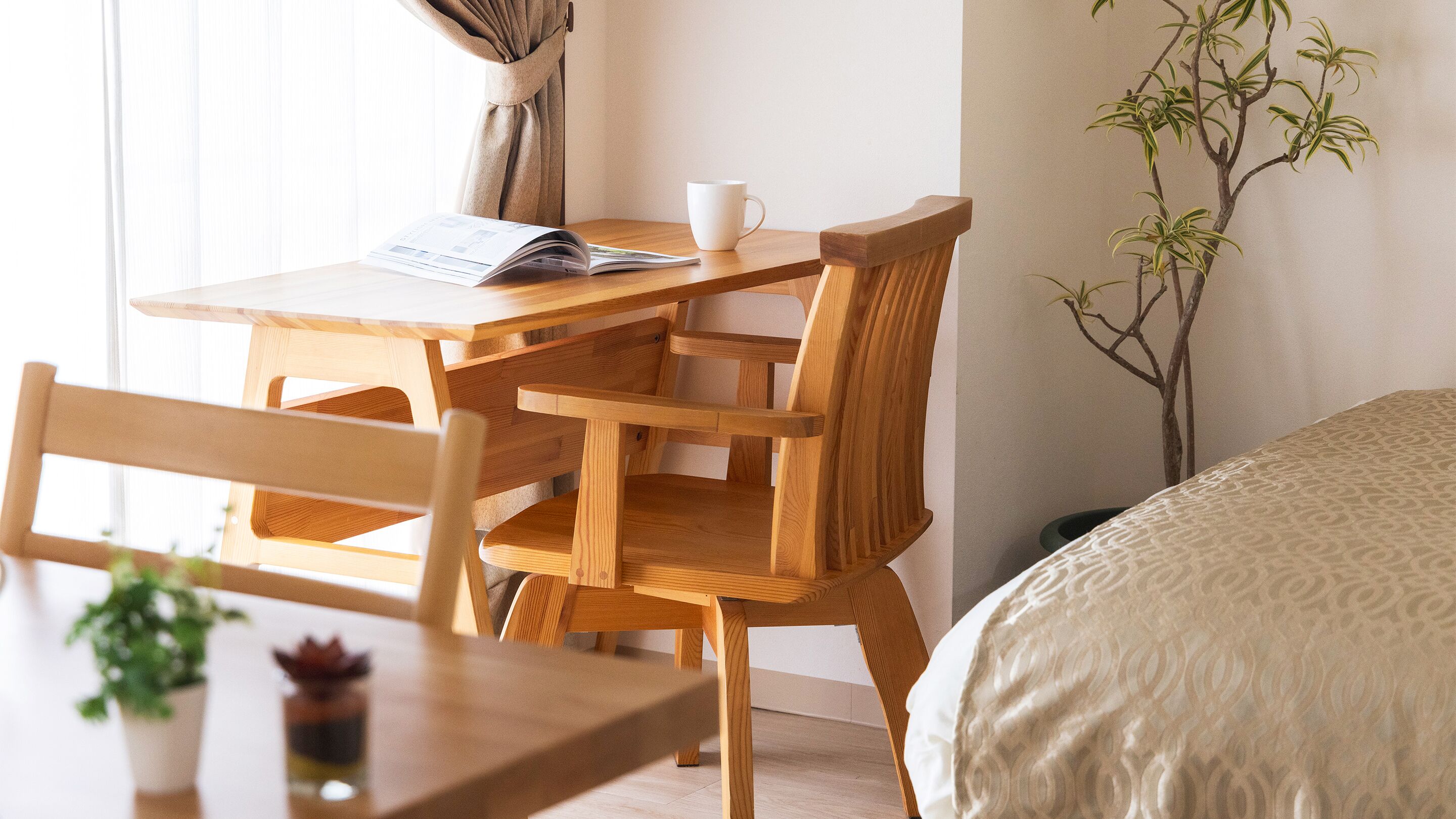 Ein Holztisch mit einer weißen Tasse und einer Zeitschrift, ein Stuhl und ein Bett mit einer gemusterten Decke.