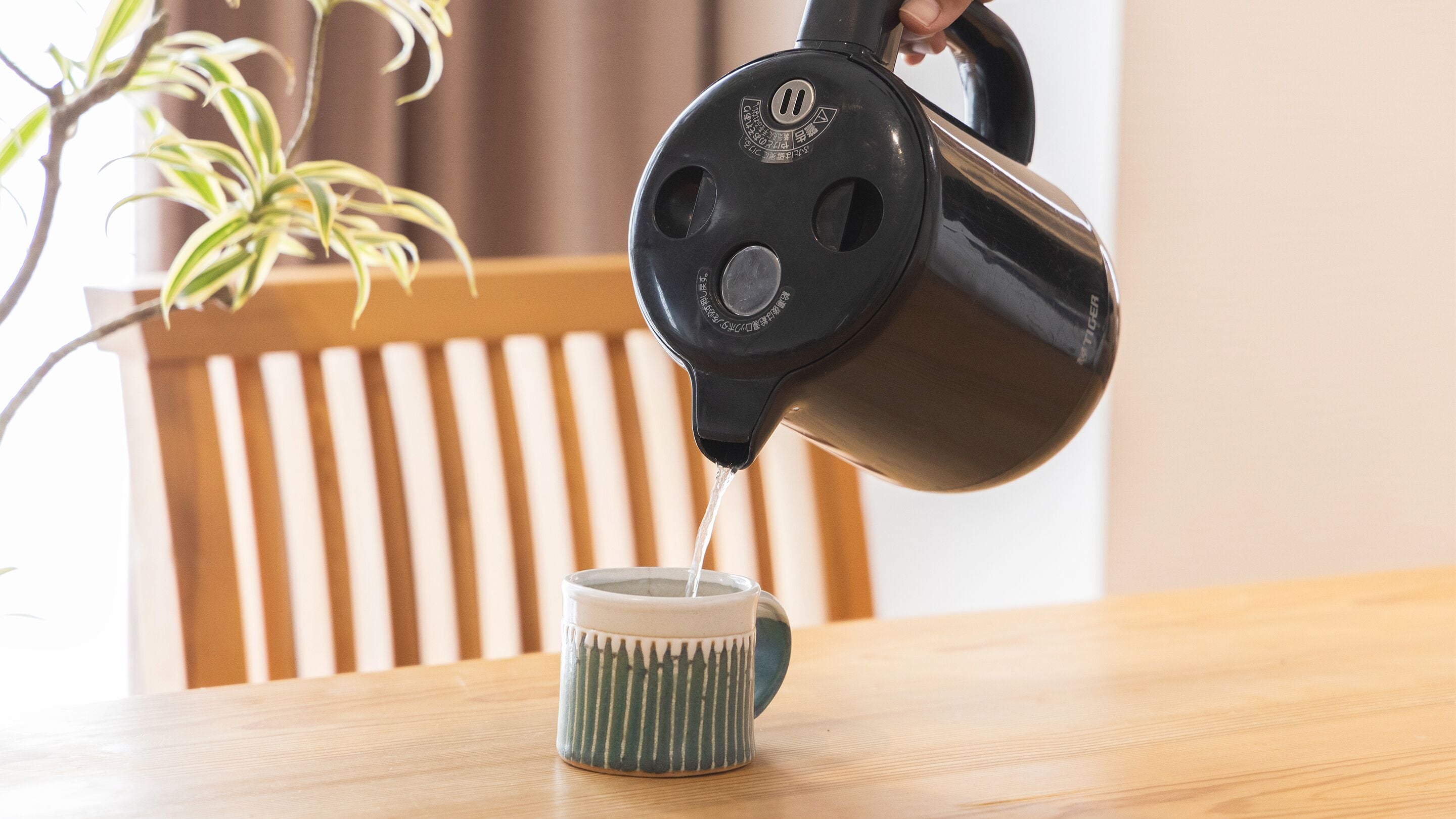 A hand pouring water from a black kettle into a green mug on a wooden table.