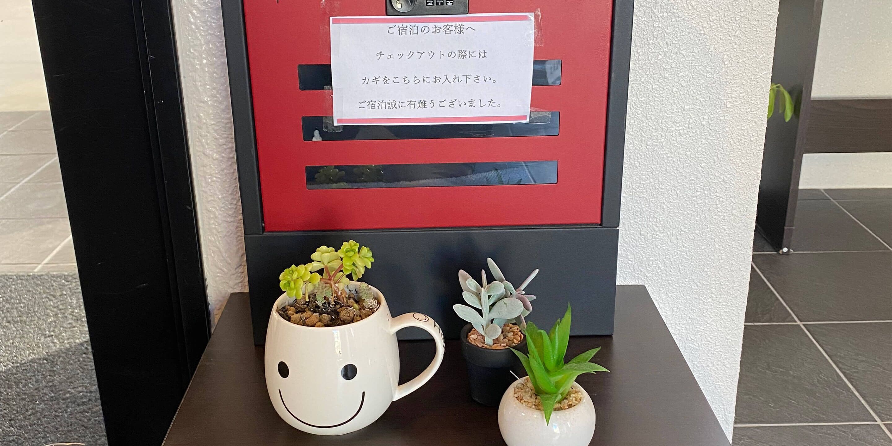 A red sign with Japanese text, a white mug with a smiley face, and two small potted plants on a table.