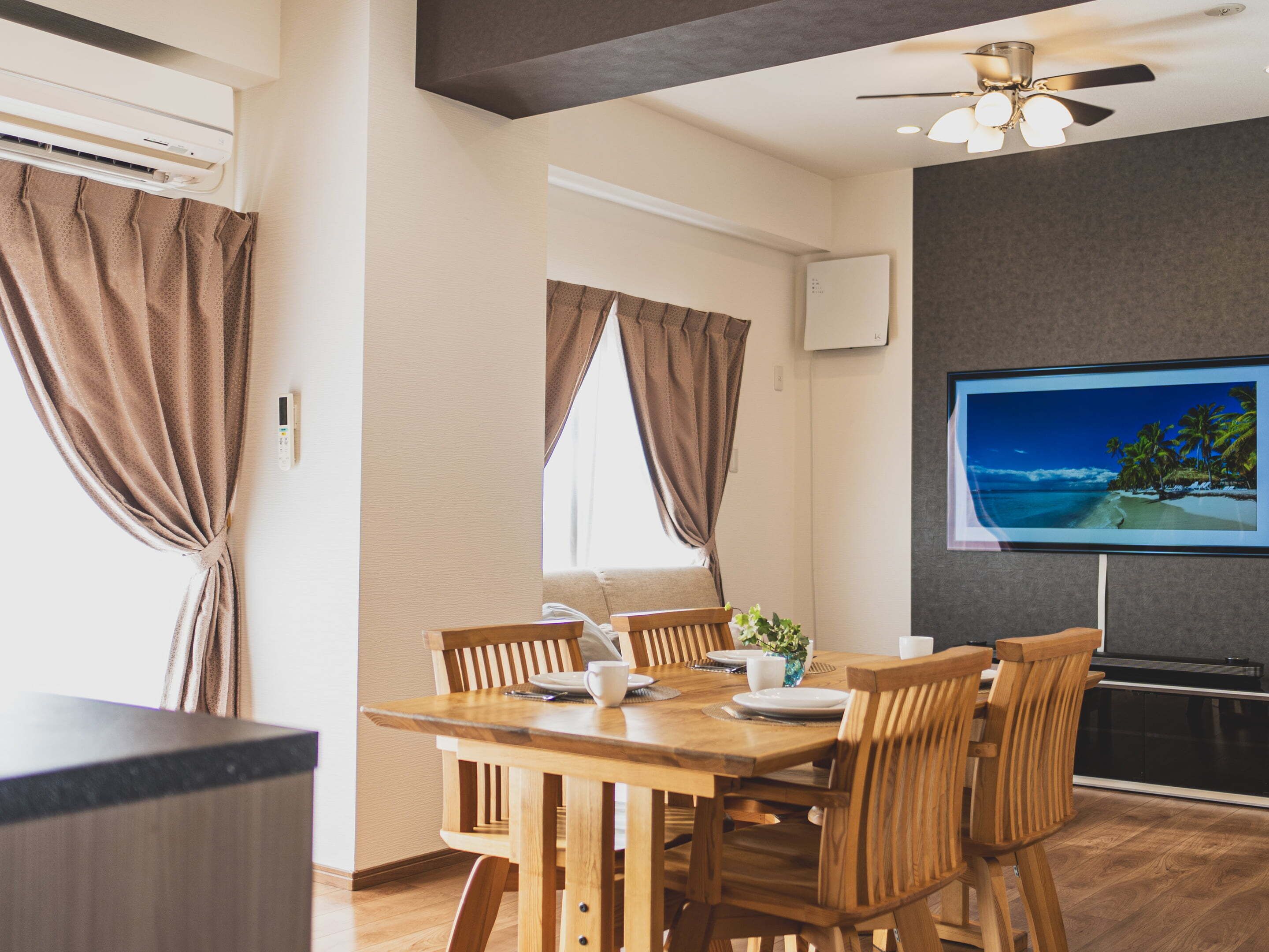 Un comedor moderno con una mesa y sillas de madera, un ventilador de techo y un televisor mostrando una escena de playa.
