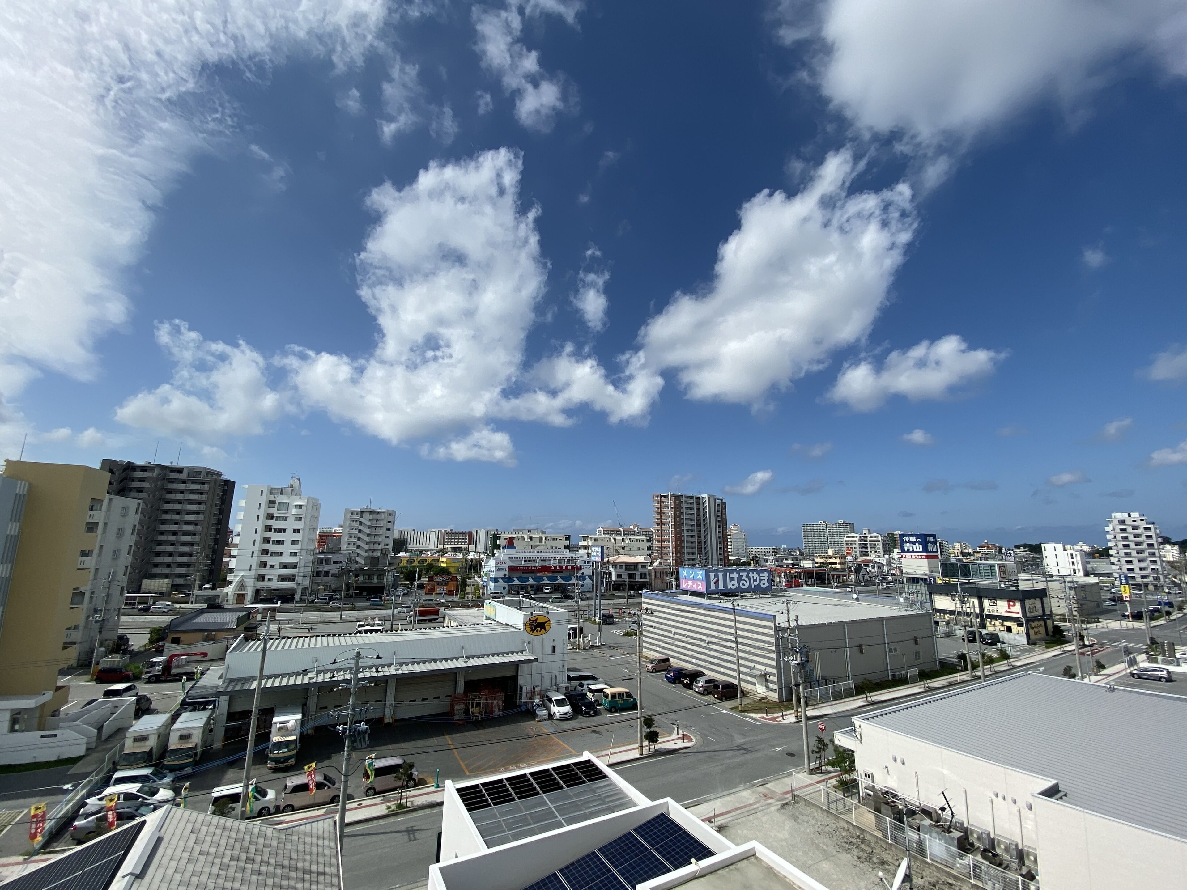 A cityscape with various buildings, including a large white structure with a flat roof and solar panels, and a smaller building with a sign in a foreign language.