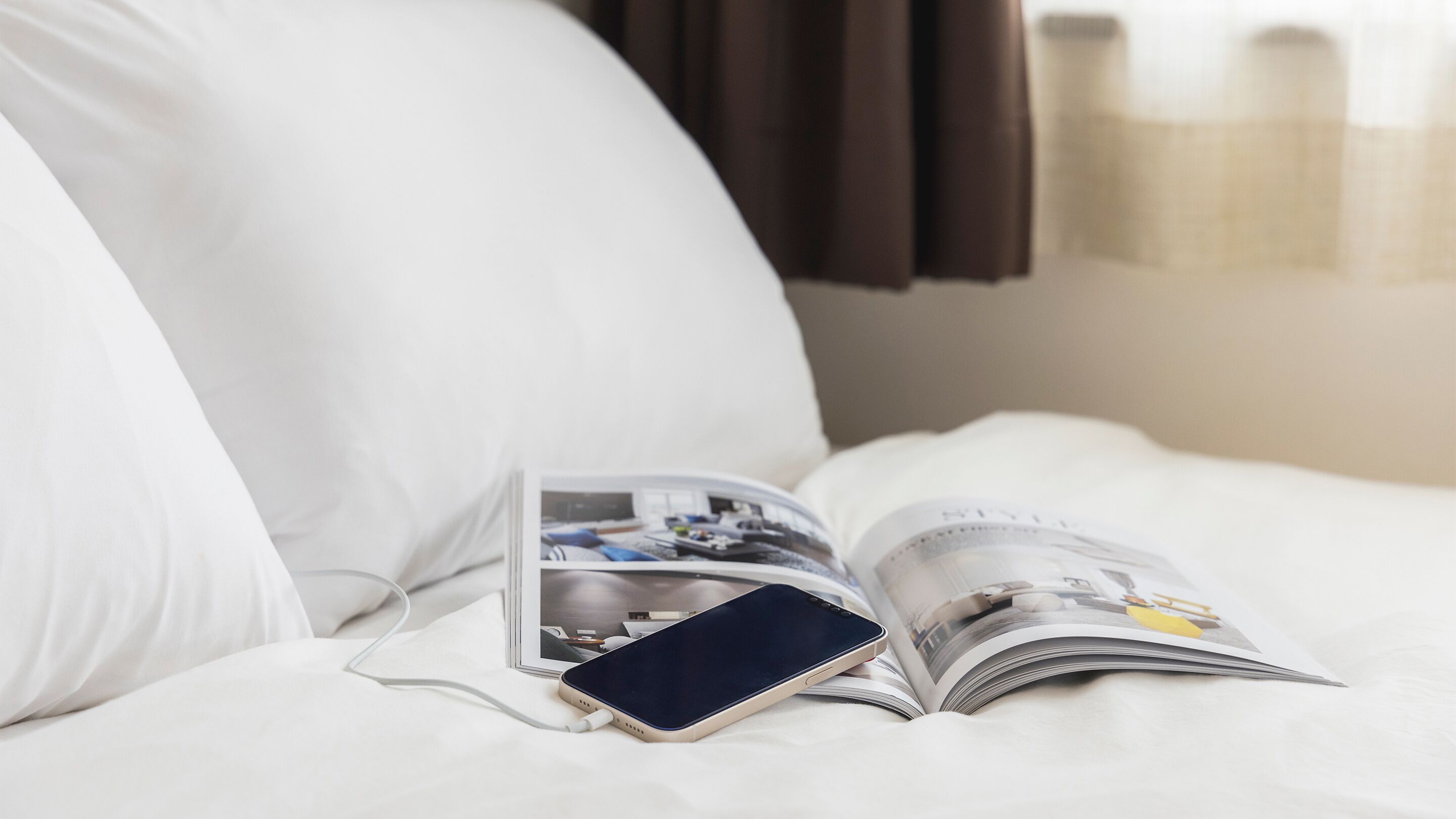 A bed with a magazine and a smartphone charging on it.