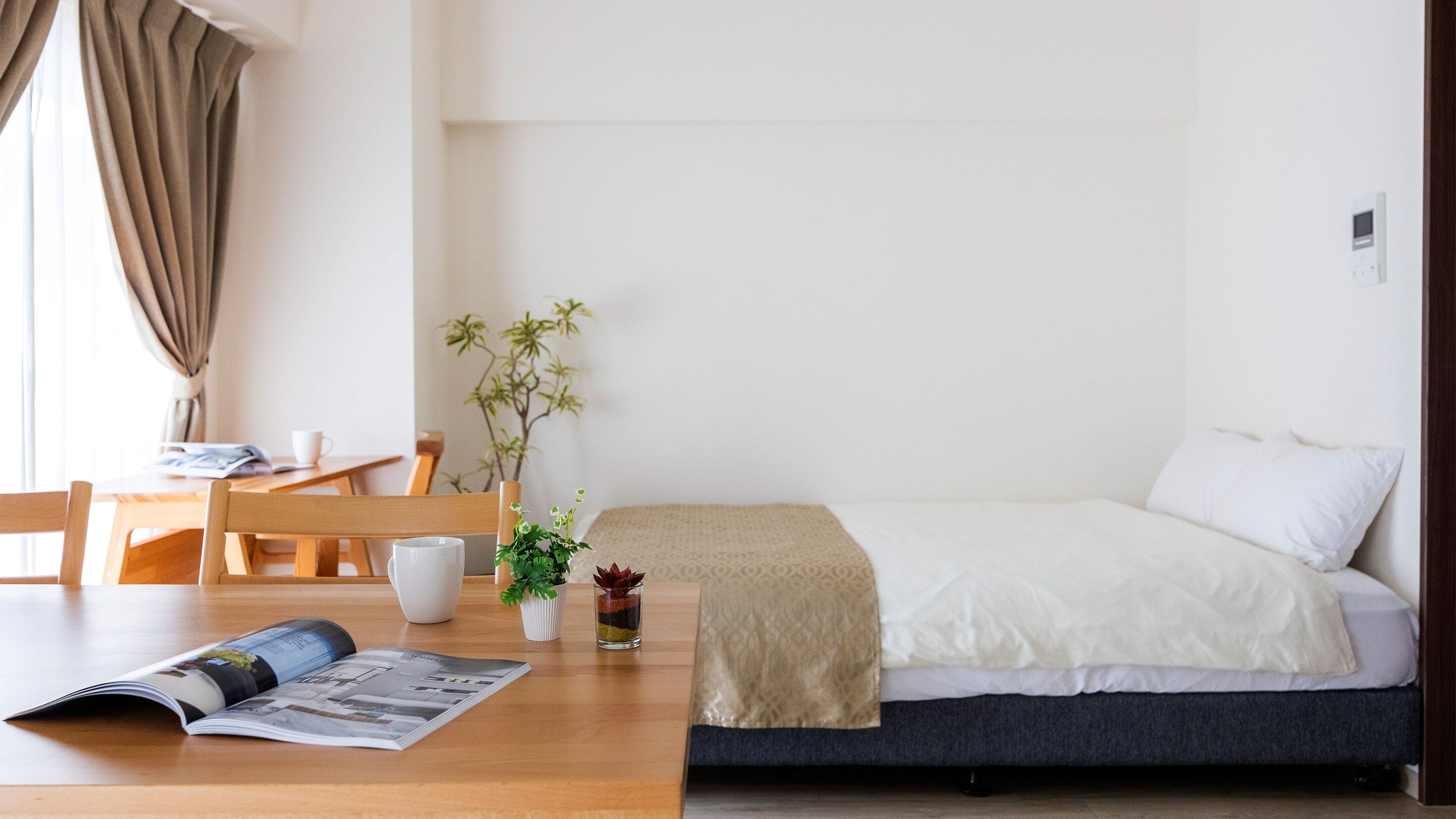 Une chambre d’hôtel minimaliste, avec un lit, une petite table sur laquelle repose un magazine, une plante et une fenêtre avec des rideaux.
