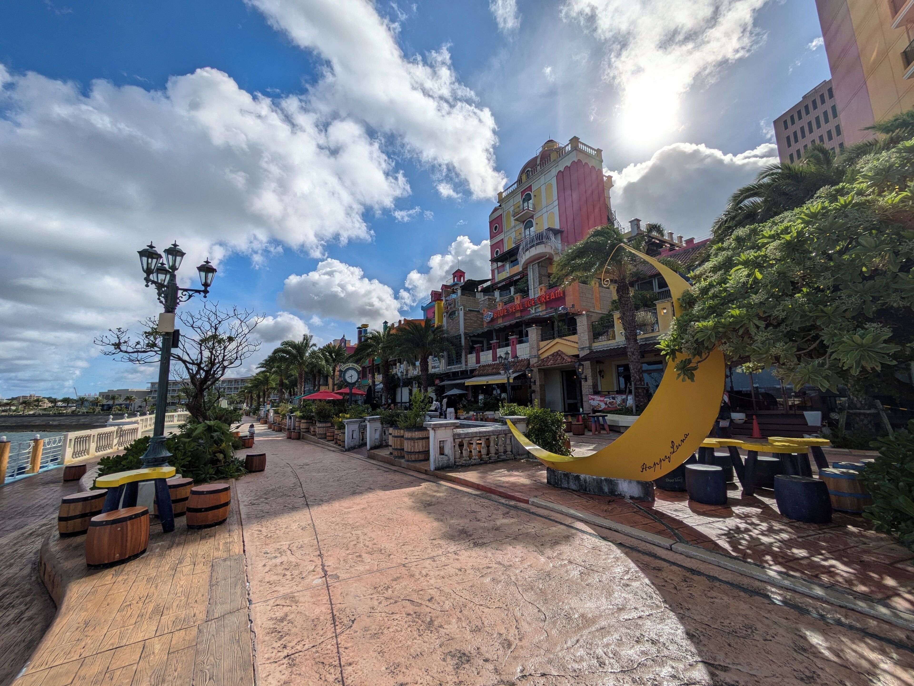 A waterfront promenade with a curved yellow bench, potted plants, and a street lamp.