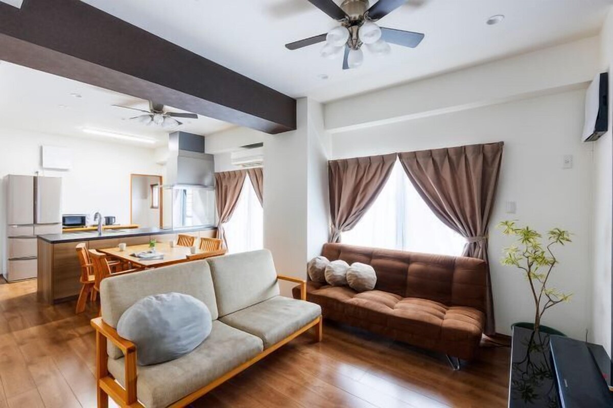Una sala moderna con un sofá, un comedor con mesa y sillas, una cocina al fondo y un ventilador de techo.