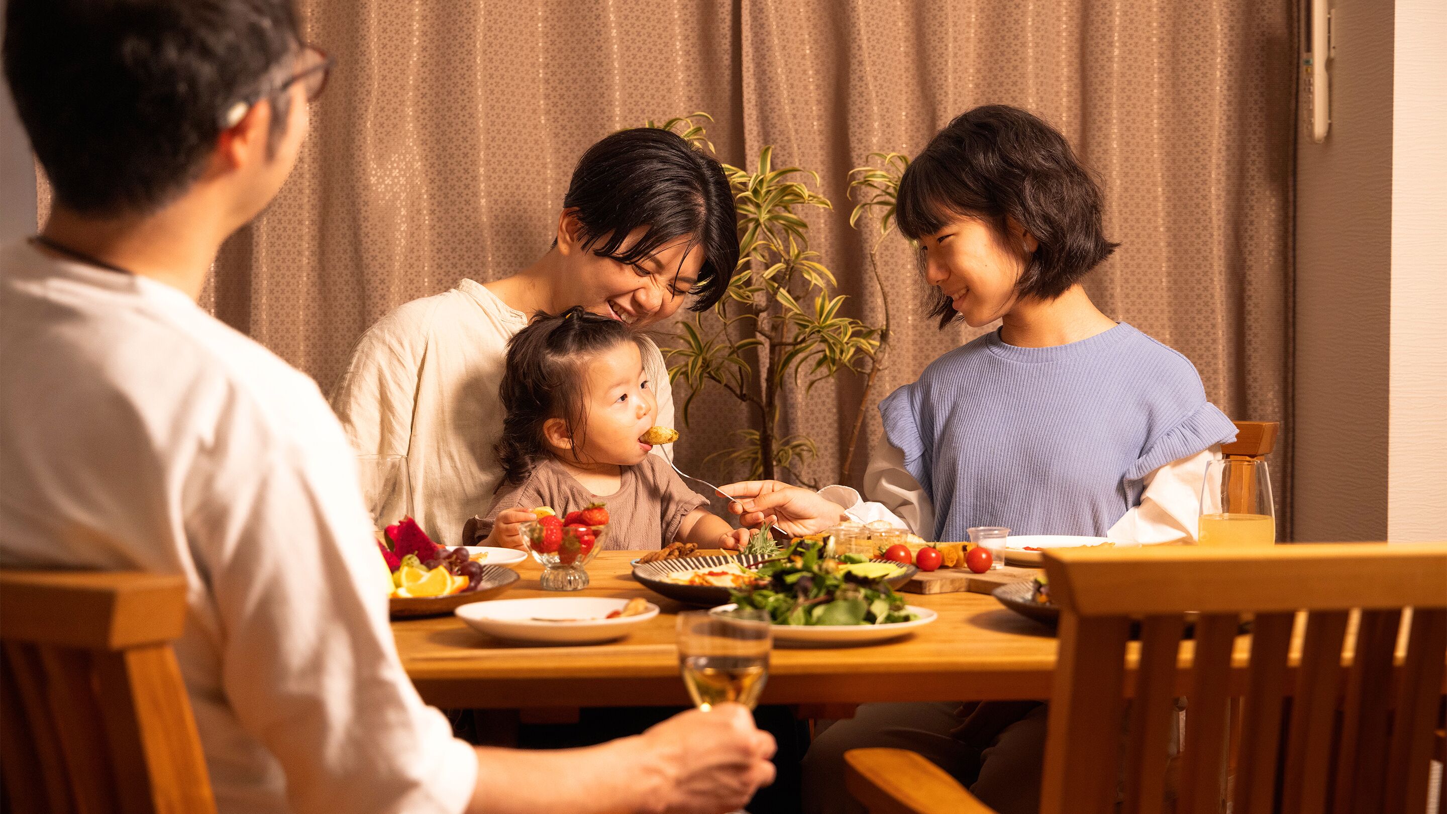 Une famille est assise autour d’une table en bois, et on peut voir différents plats, un verre de vin et une plante en pot à l’arrière-plan.