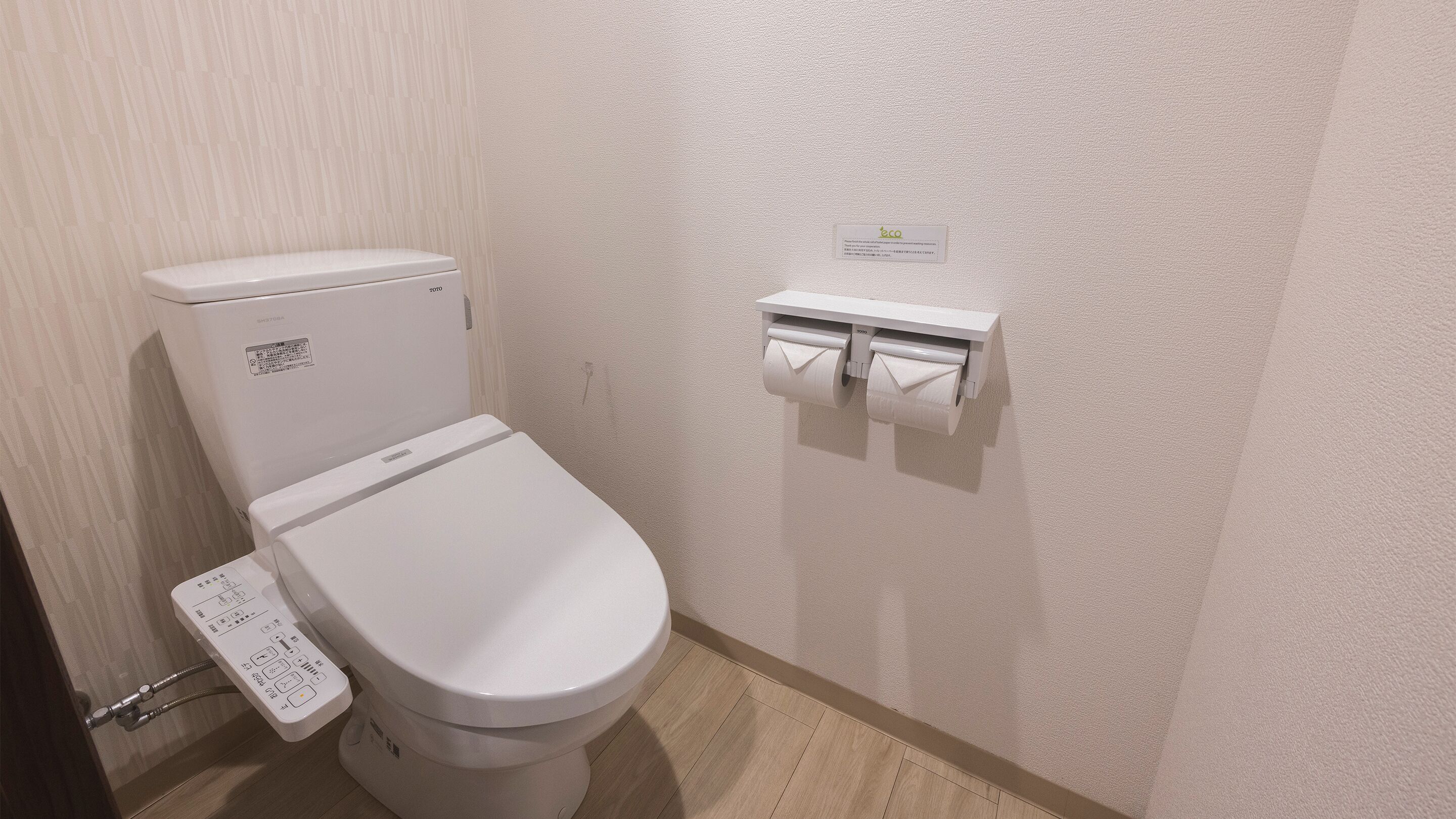 Un WC standard bianco con l'etichetta del marchio Toto, un portasciugamani con due rotoli di carta igienica e un piccolo cartello appeso al muro.