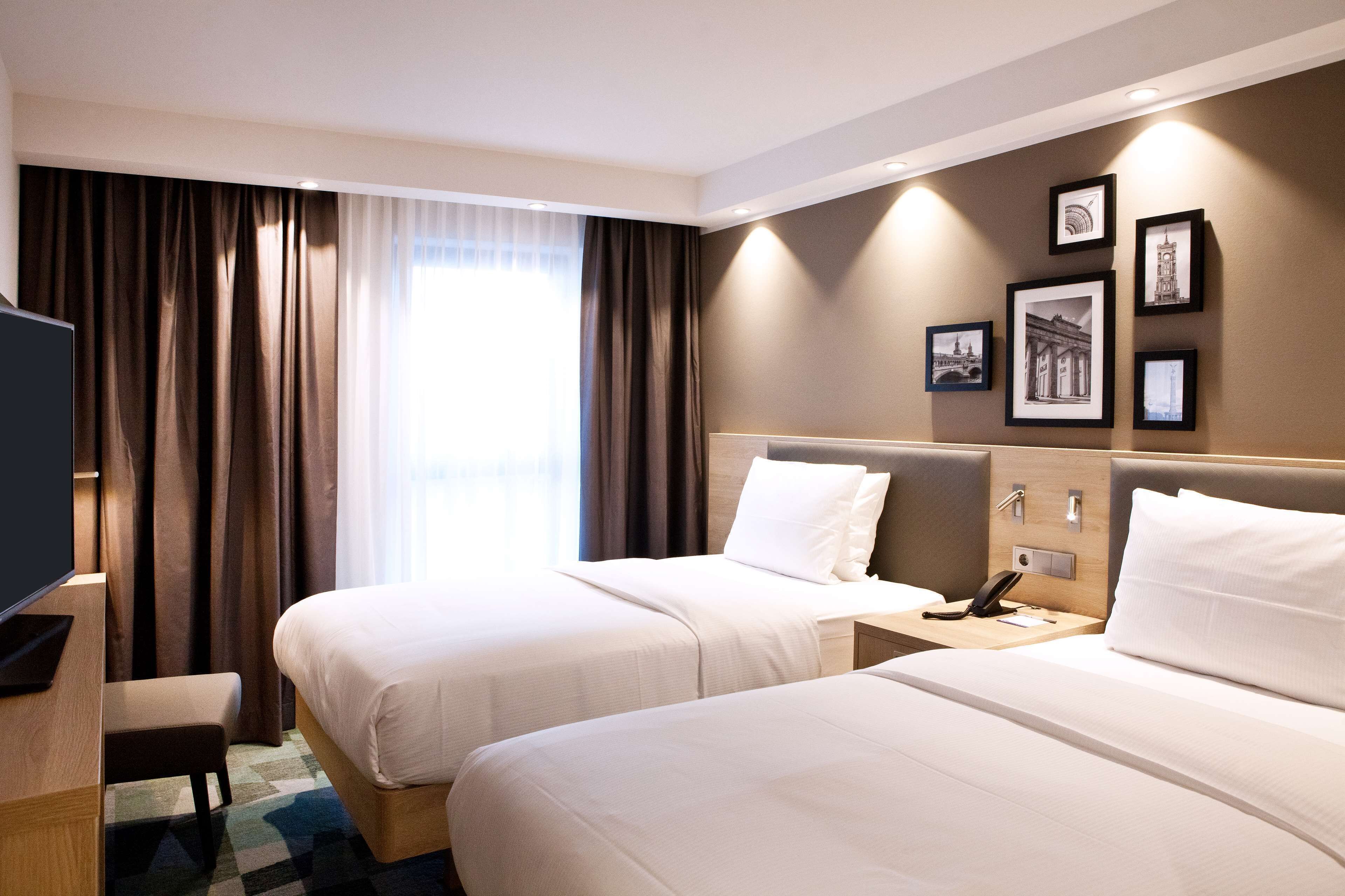 A hotel room with two beds, a desk, a TV, and a wall with framed pictures.