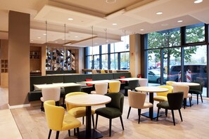 Un café moderne avec des tables et des chaises rondes, un coin salon avec un canapé vert et une grande fenêtre donnant sur la rue.