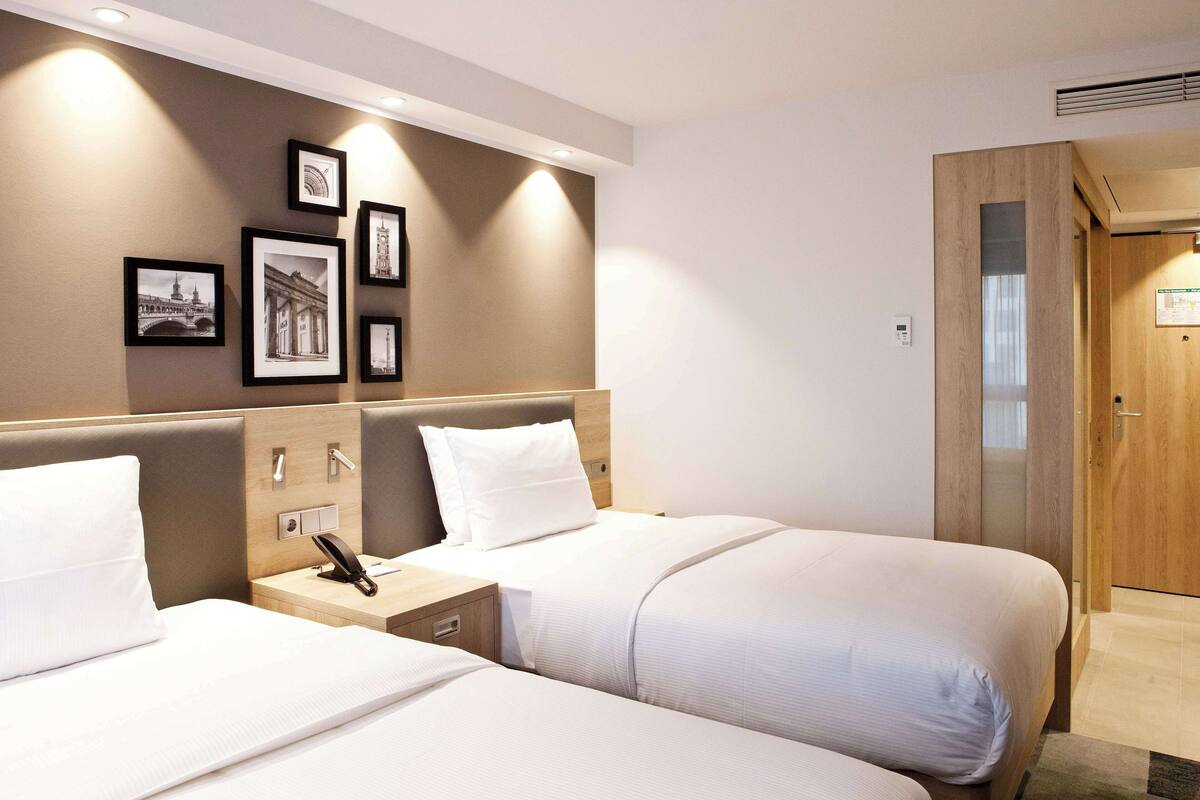 Habitación de hotel con dos camas, una mesita de noche con teléfono, luz de pared e imágenes enmarcadas.