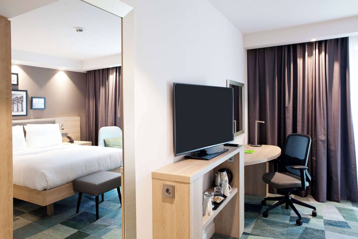 Habitación de hotel con cama, escritorio con computadora, silla, televisor y ventana con cortinas.