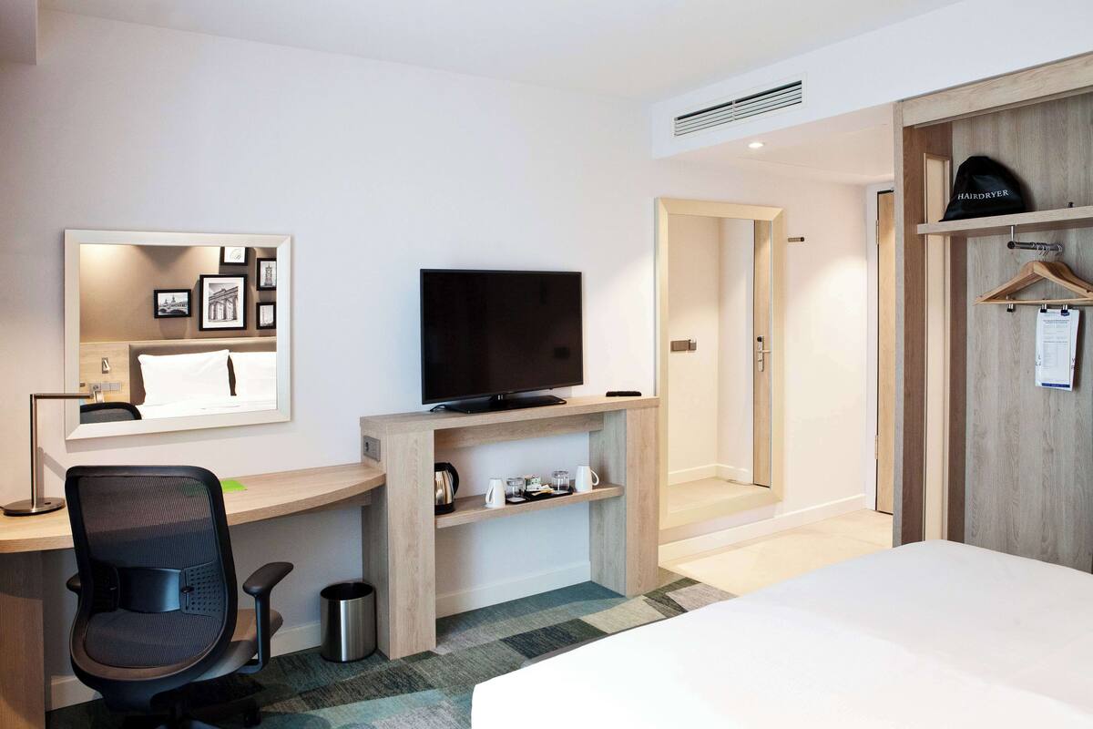 Una habitación de hotel con cama, escritorio con televisor, silla, espejo y armario.