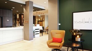 Un moderno vestíbulo de hotel con una mesa de recepción, un área de descanso con una silla y una mesa, y una obra de arte con un cuadro de la ciudad en la pared.