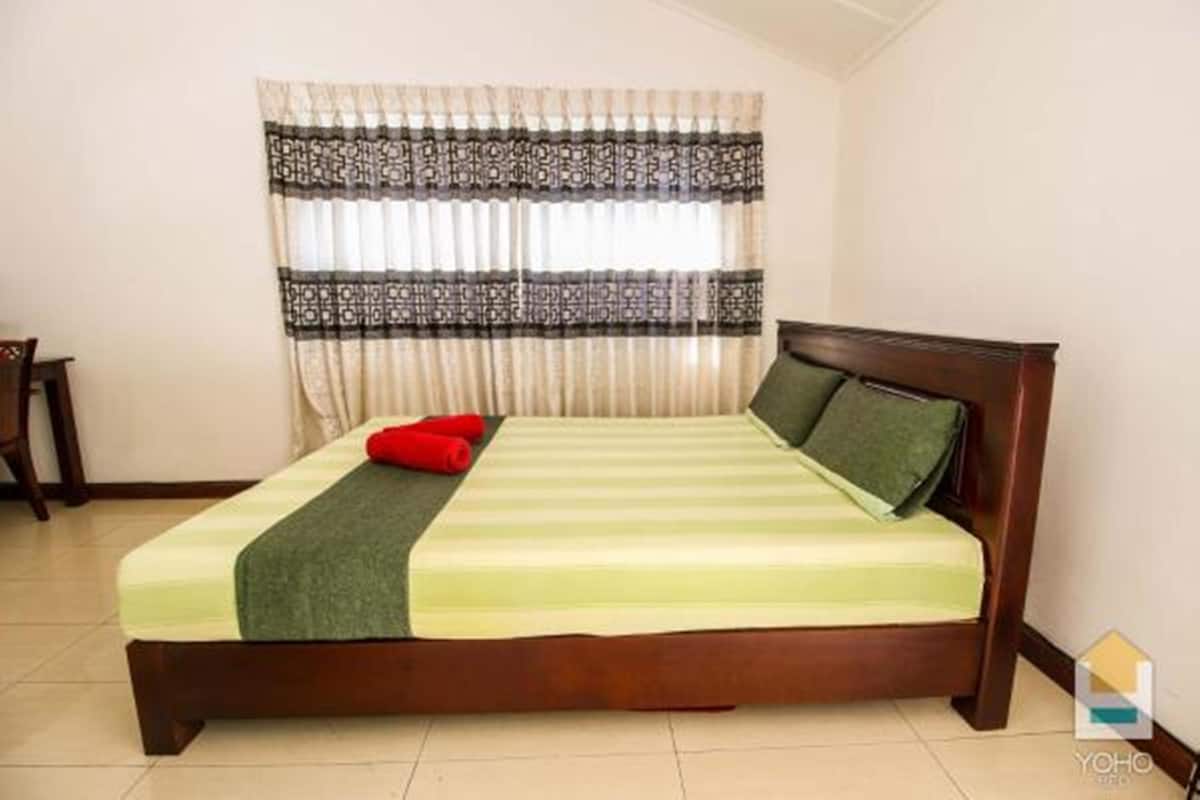 Un lit bien fait, recouvert d’une couvre-lit à rayures vertes et blanches, avec un oreiller rouge et un oreiller vert. Un rideau à motifs est accroché au-dessus du lit.