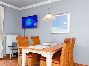 Un coin repas avec une table en bois et des chaises en osier, un téléviseur fixé au mur et une fenêtre donnant sur un beau paysage.