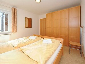 Une chambre avec deux lits, une armoire en bois, un petit tabouret et une fenêtre avec des rideaux.