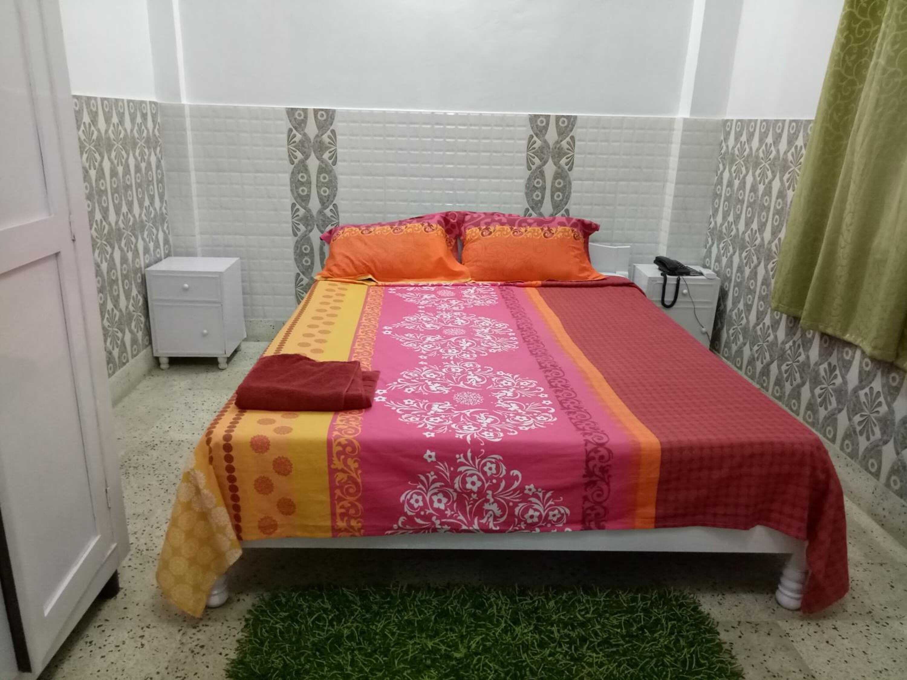 A neatly made bed with a patterned bedspread, a bedside table, and a green rug.