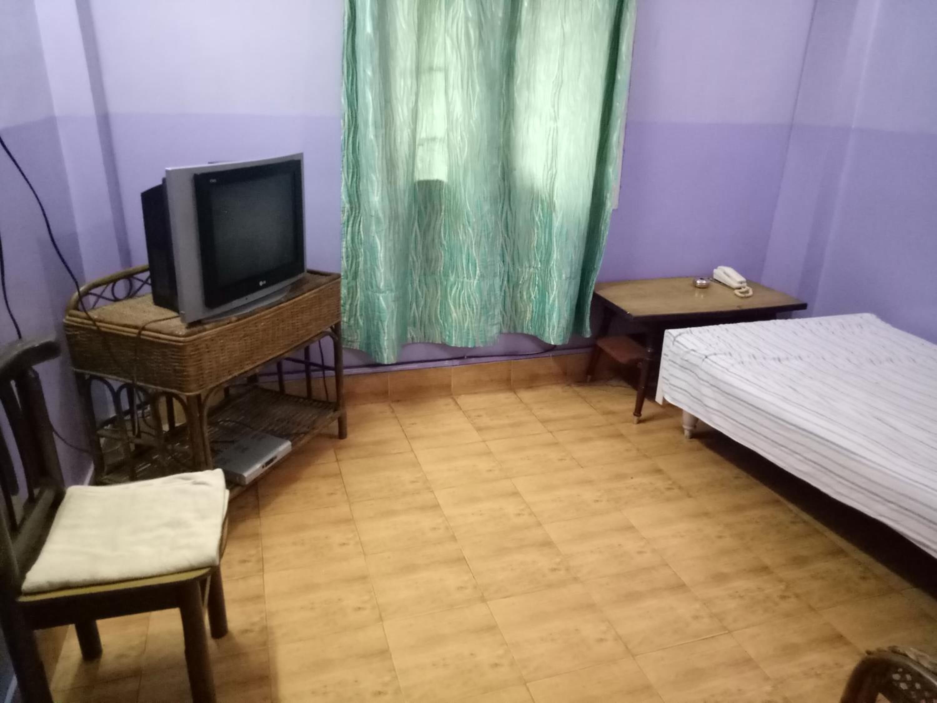 A small room with a television on a stand, a chair, a bed, a small table, and a curtain.