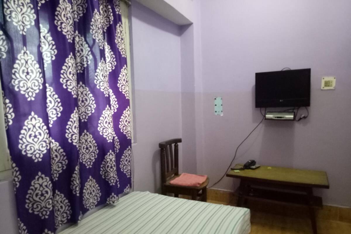 Una habitación con cama, una silla, un escritorio, televisor montado en la pared y una cortina morada con estampado.