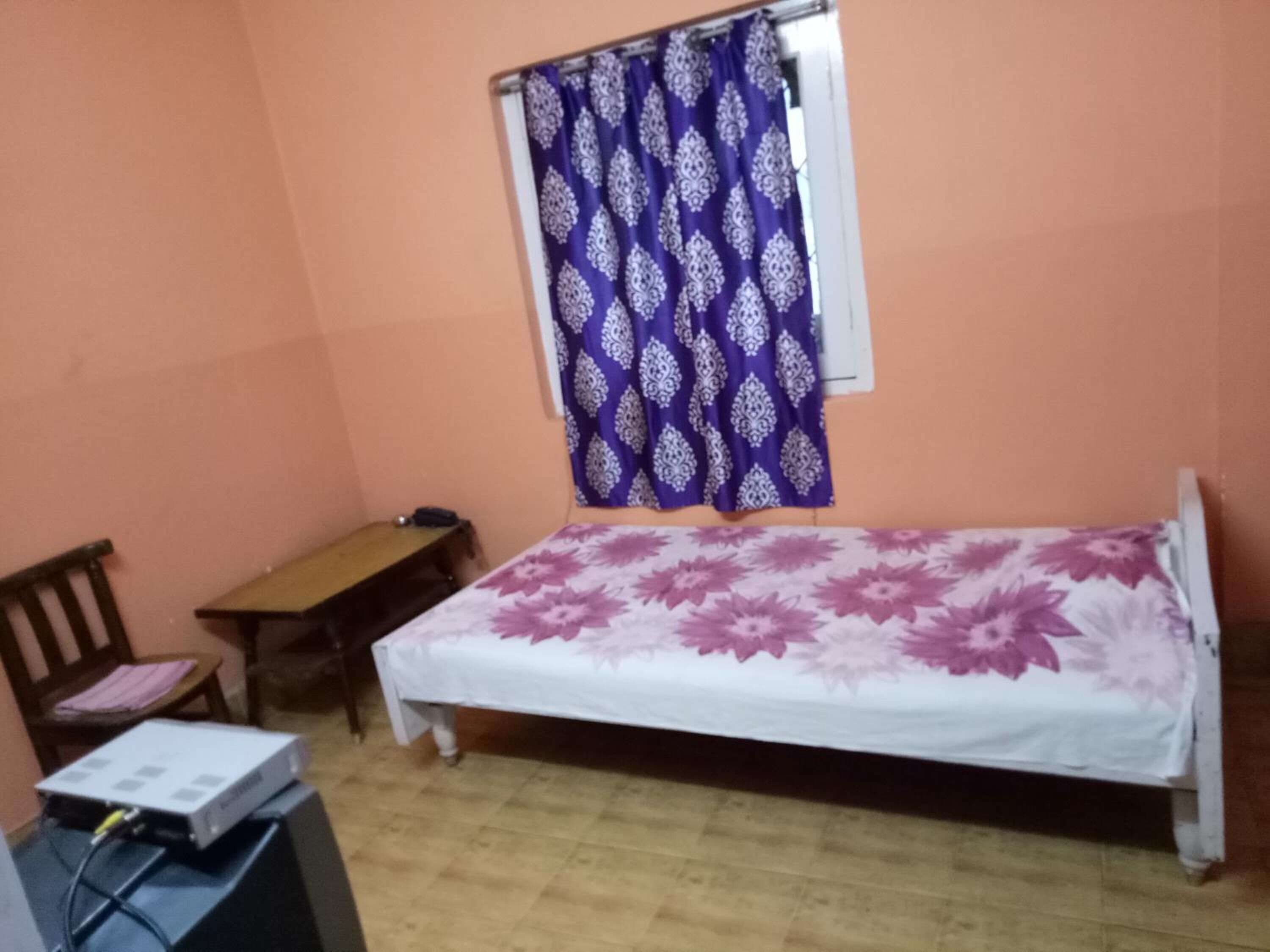A small bedroom with a single bed, a wooden desk, a chair, and a window with a patterned curtain.