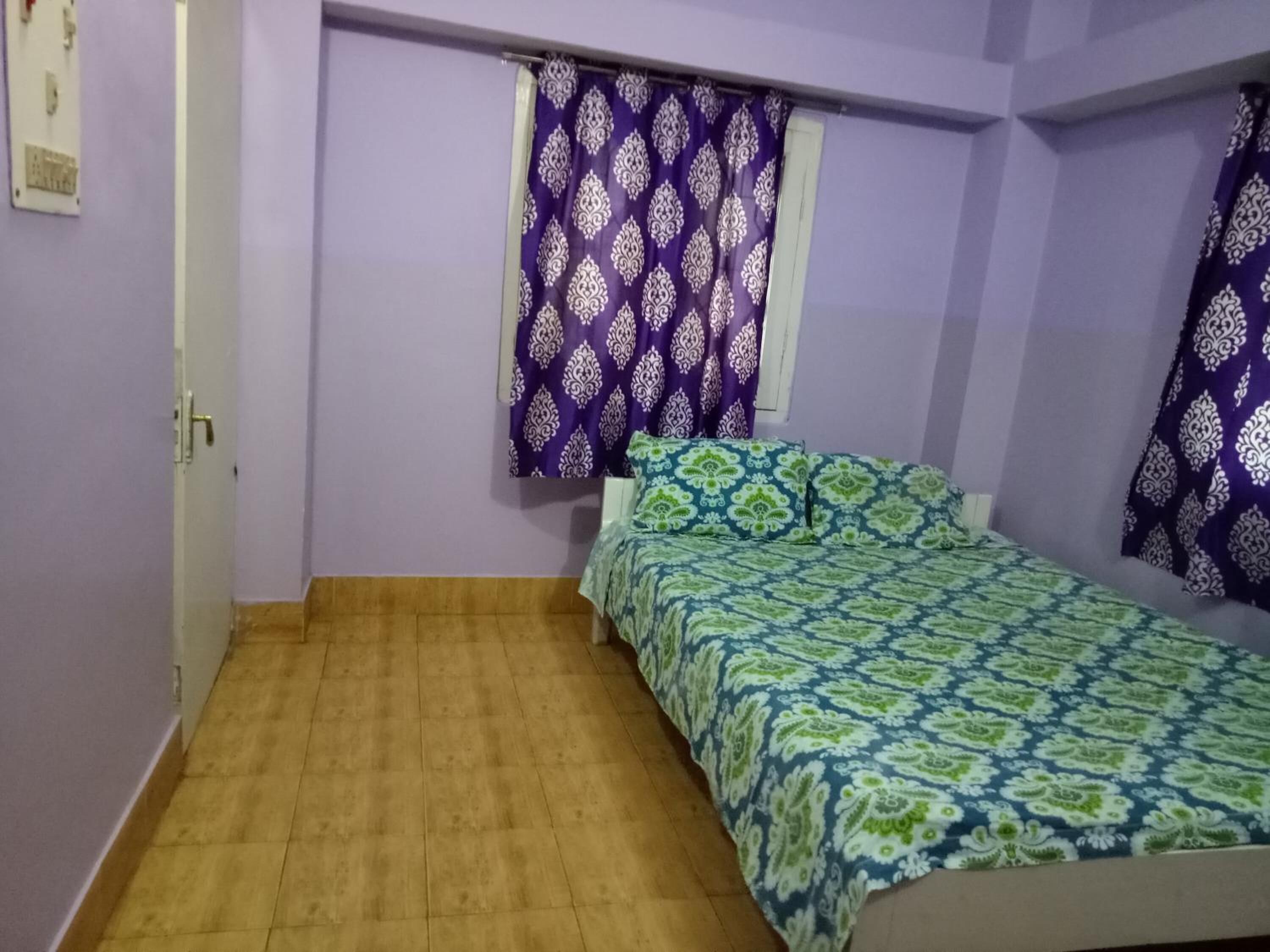 A small, single-bed room with a wooden floor, purple walls, and patterned curtains.