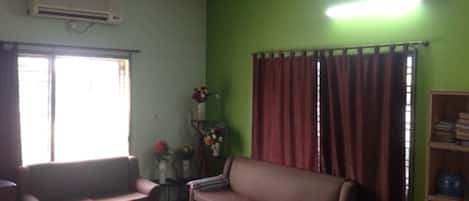 A living room with a sofa, a coffee table, a ceiling fan, an air conditioning unit, a window with curtains, and a bookshelf.